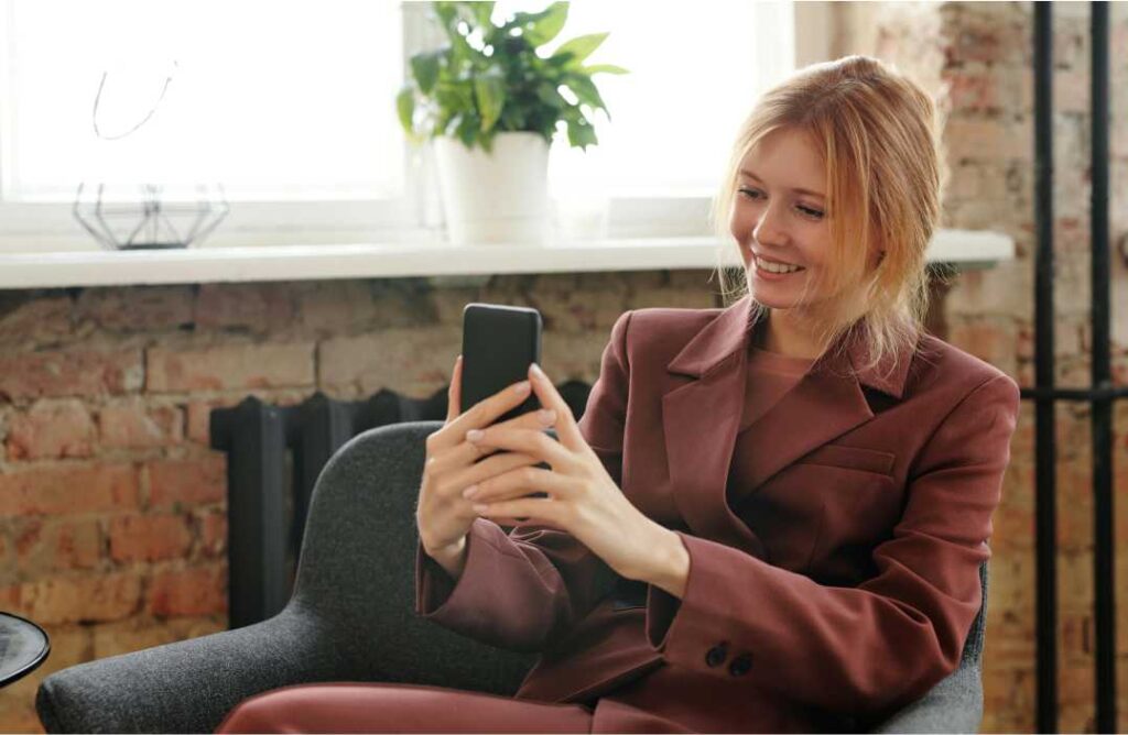 Nasmijana poslovna žena u elegantnom smeđem saku sjedi u uredu i na mobitelu prati inspirativne žene na LinkedInu.