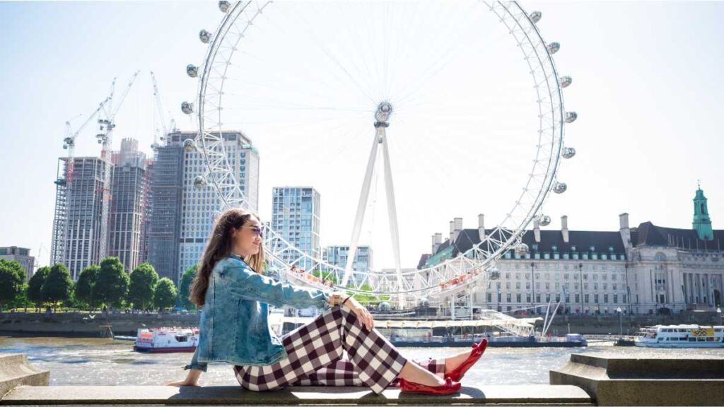 Djevojka sjedi uz rijeku Temzu s pogledom na London Eye, dok istražuje najpoznatije atrakcije u Londonu.