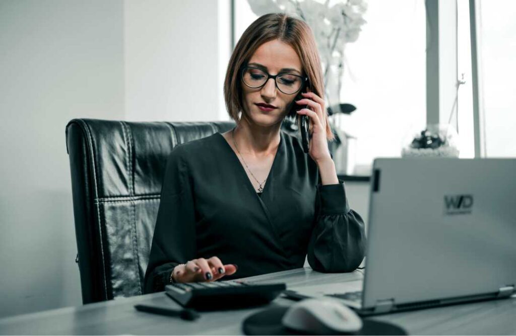Žena u uredu koja istovremeno telefonira i radi na računalu kao prikaz profila žene robota, stanja gdje nedostaju humor i emocionalna inteligencija.