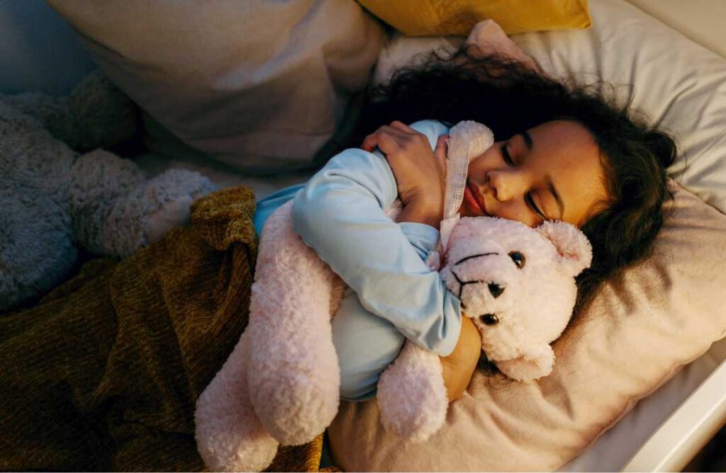 Priča o nadi: Djevojčica crne kovrčave kose spava grleći plišanog medvjedića kao simbol sigurnosti.