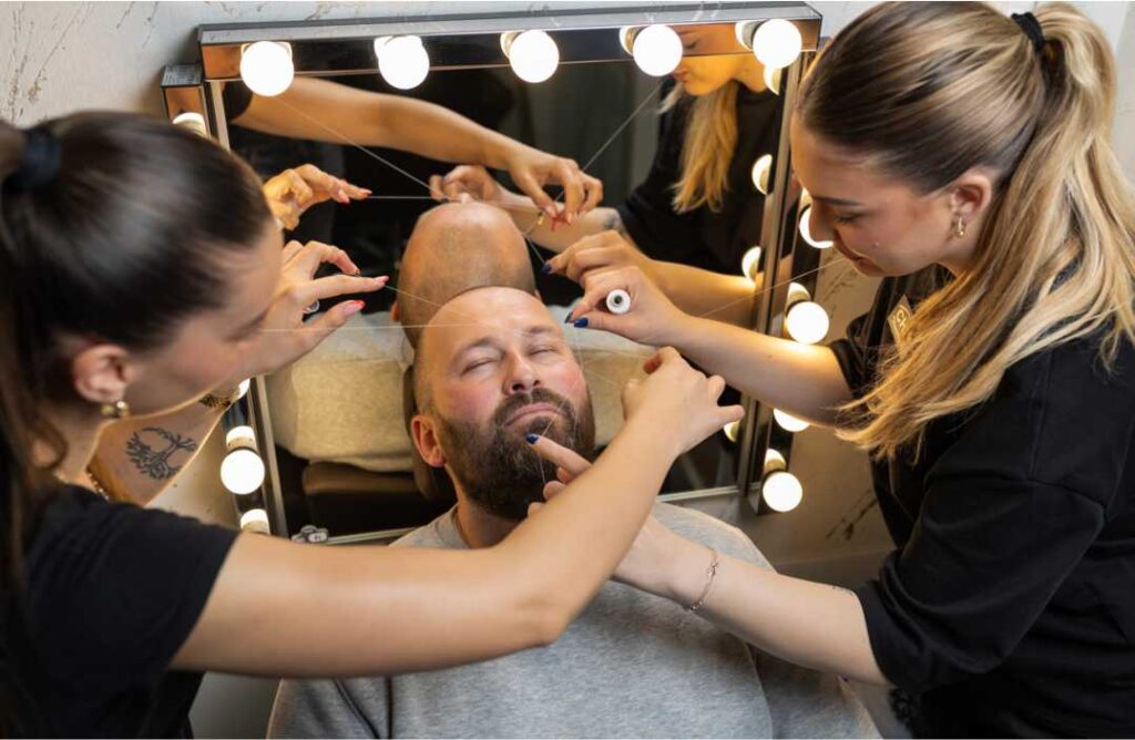 Muškarac s punom bradom koji mirno sjedi u stolici, dok mu tri žene profesionalno oblikuju bradu koncem (threading). Slika je snimljena iz ptičje perspektive i prikazuje ih ispred ogledala sa žaruljama. Žene koriste fine konce za precizno oblikovanje brade i obrva.