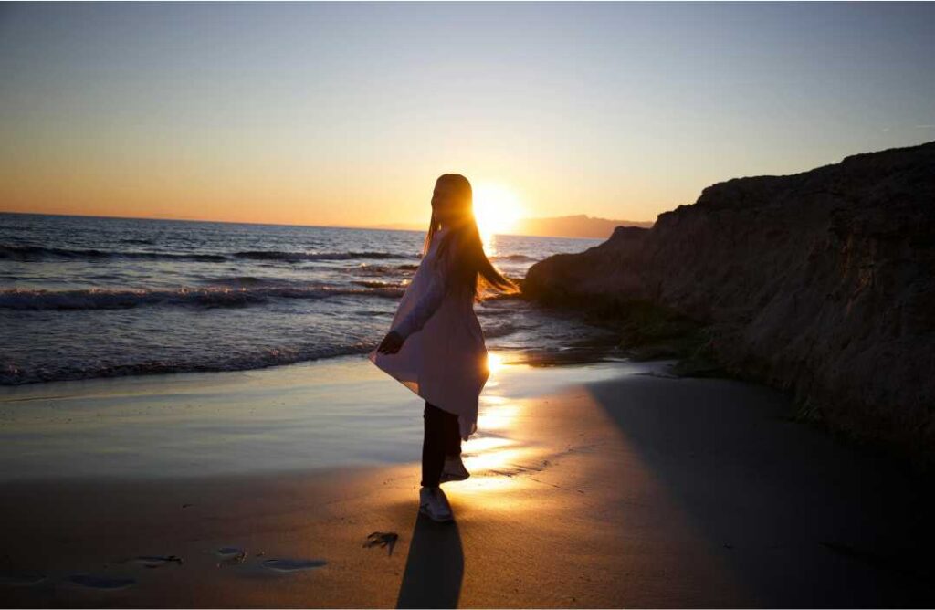 Osoba na pješčanoj plaži u dinamičnom, plesnom pokretu koji utjelovljuje jedinstvo duha i tijela i holistički ritam života, okupana zlatnom svjetlošću zalaska sunca.