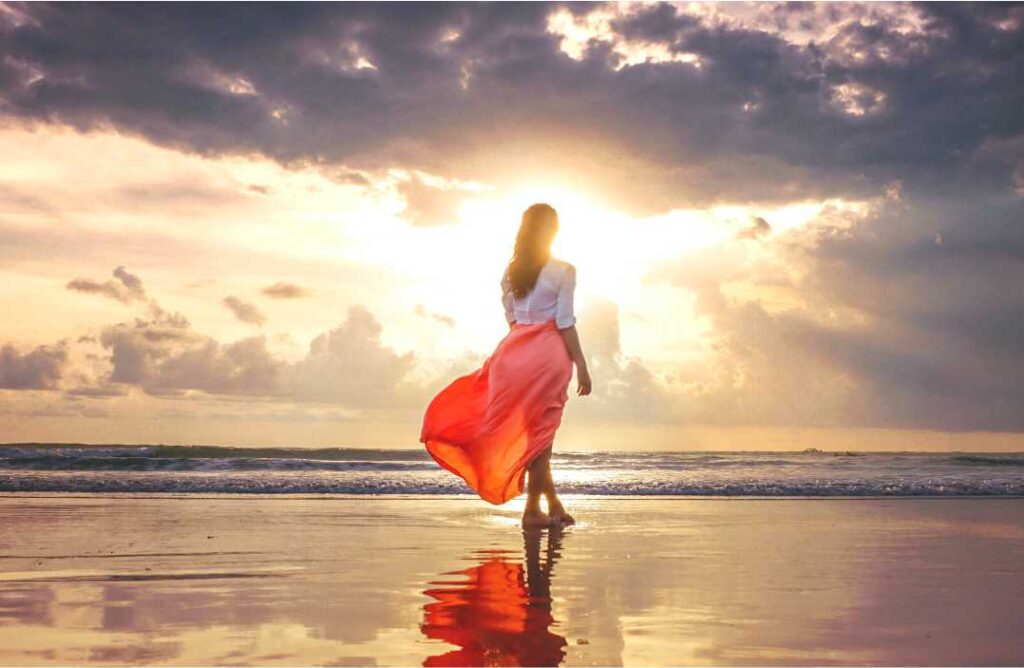 Mlada žena u crvenoj suknji hoda prema suncu na plaži – vizualna metafora za akciju i hrabrost tranzita Mars u Ovnu.