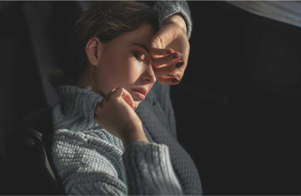 Mlada žena u sivom džemperu drži ruku na čelu u zamračenoj sobi, ilustrirajući mentalnu napetost i potrebu za povlačenjem, što su ključne teme tranzita Uran u Blizancima.