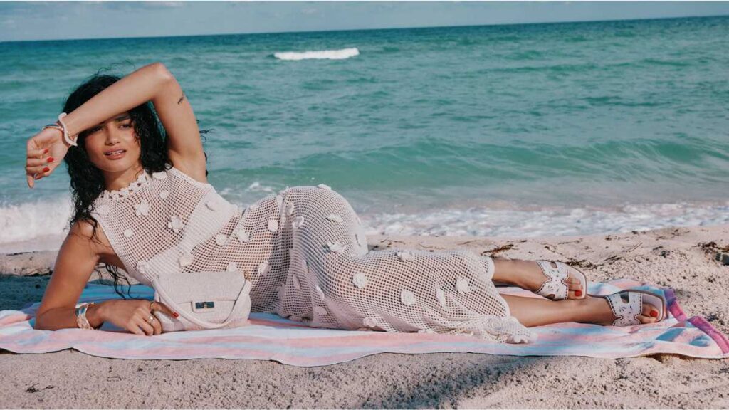 Model na plaži predstavlja modele iz ponude Aldo ljetna kolekcija, noseći bijelu pletenu haljinu, ravne natikače s izrezima i pripadajuću torbicu.