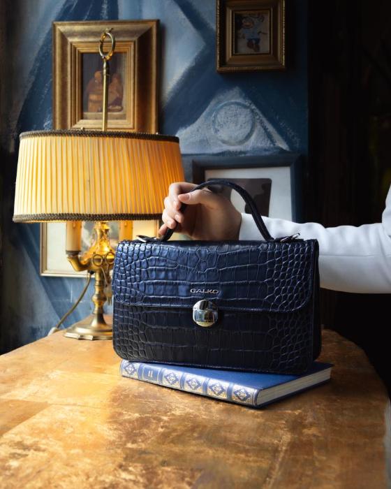 Galko torbe u klasičnom stilu: ženska ruka drži elegantnu crnu kožnu torbu s upečatljivom krokodilskom teksturom, položenu na staru plavu knjigu na drvenom stolu pokraj vintage stolne lampe s toplim žutim svjetlom.