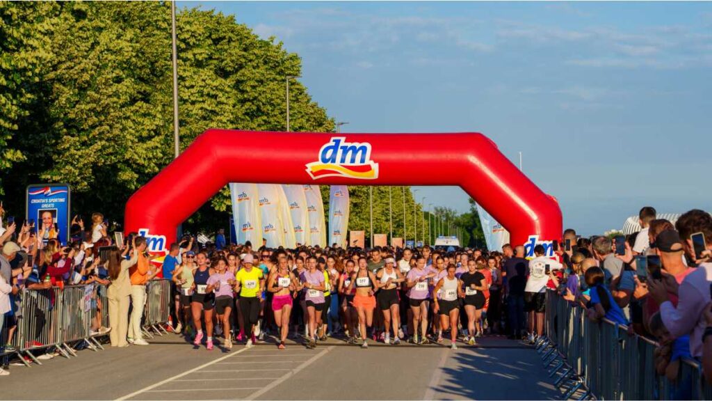 dm ženska utrka okupila više od 9.000 sudionika i osigurala 18.002 eura za Udrugu P.I.N.K. – life Start utrke dm ženska utrka Jarun 2026 s velikim brojem trkačica koje prolaze ispod crvenog dm luka na napuhavanje, okružene publikom koja navija iza ograda.