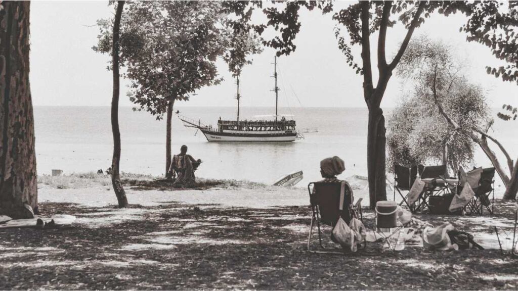 Katarina Solomun: Žaljenje za prošlim vremenima Crno-bijela vintage fotografija koja dočarava bezbrižno ljetovanje u bivšoj državi: ljudi se odmaraju u debeloj hladovini drveća na plaži, dok je u pozadini na mirnom moru usidren stari jedrenjak.