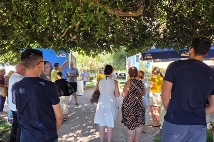 Građani okupljeni u hladu drveća čekaju na besplatni dermatološki pregled na dm-ovom punktu na otvorenom.