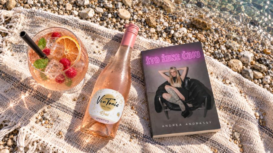 Knjiga Andree Andrassy „Što žene čuju“, boca pića VinTonic Rosé i čaša koktela na prostirci na šljunčanoj plaži.