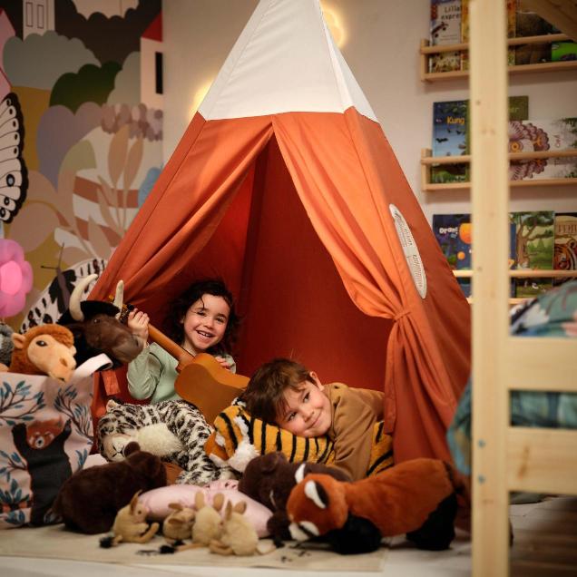 Dvoje nasmijane djece leži u narančastom IKEA dječjem šatoru (tipiju), okruženi mnoštvom mekanih plišanih igračaka životinja s Himalaje (snježni leopard, crvena panda, jak, tigar) iz ALPTALL kolekcije.