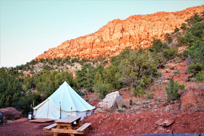 Bijeli glamping šator s drvenim stolom za piknik u Zion View Campingu, smješten u podnožju crvenih stijena i pustinjskog krajolika Utaha. Ovakvi neobični smještaji za odmor omogućuju boravak u prirodi uz potpunu udobnost.