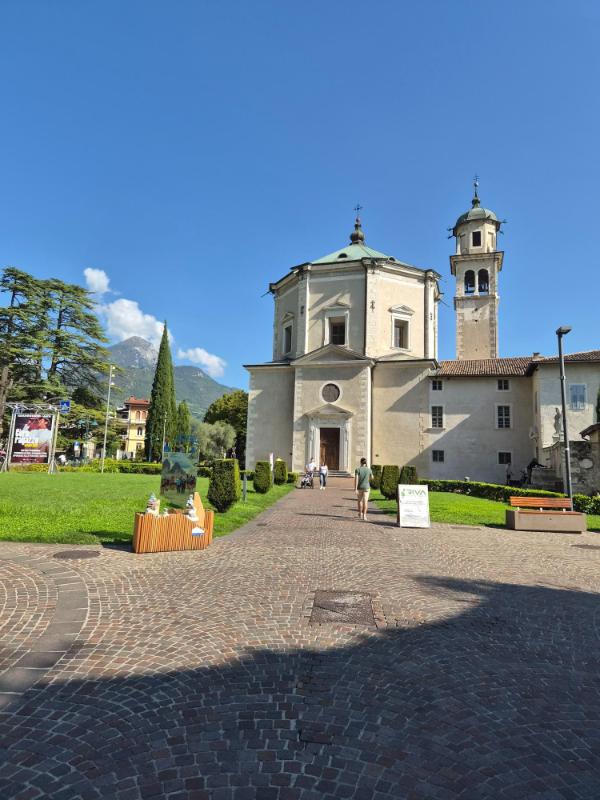 Eksterijer barokne crkve Chiesa dell'Inviolata s visokim zvonikom i zelenim prilaznim parkom u gradu Riva del Garda.