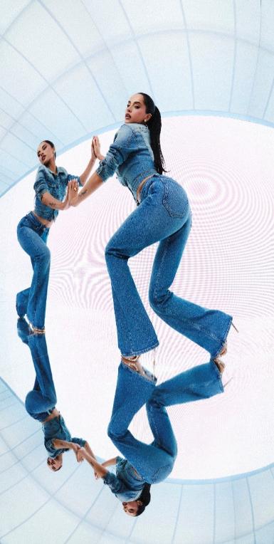 Umjetnička modna fotografija Becky G u GUESS B Jean trapericama niskog struka i traper jakni, s dinamičnim zrcalnim odrazom na podu, u modernom studijskom okruženju.
