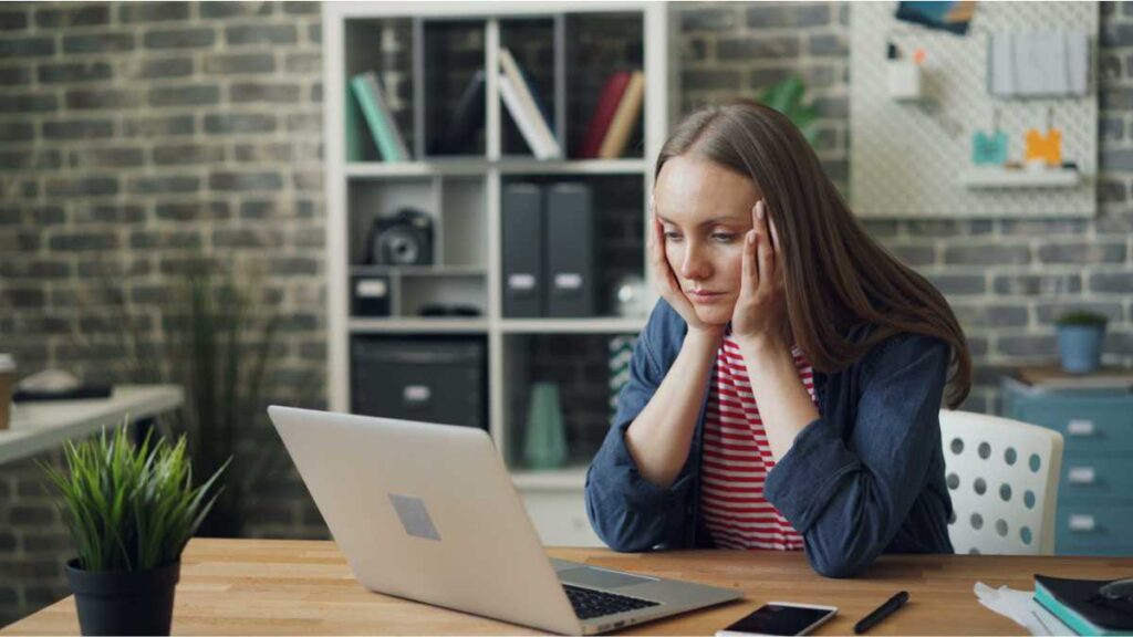 Umorna žena sjedi za radnim stolom ispred laptopa i razmišlja kako se natjerati na vježbanje nakon posla.