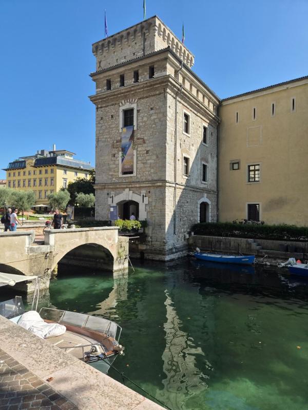 Srednjovjekovna utvrda Rocca di Riva u gradu Riva del Garda s kamenim mostom i odrazom u tirkiznom kanalu s brodicama.