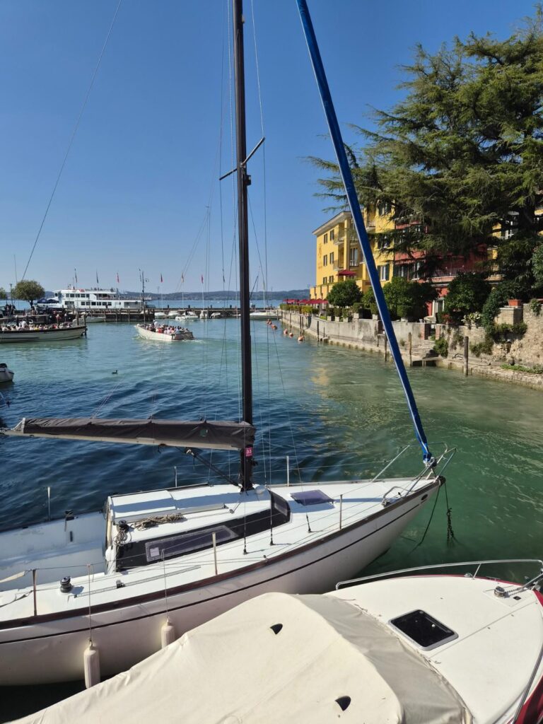 Jezero Garda, pogled na tirkizni akvatorij i luku s jedrilicama, turističkim gliserima i obalnim zgradama.