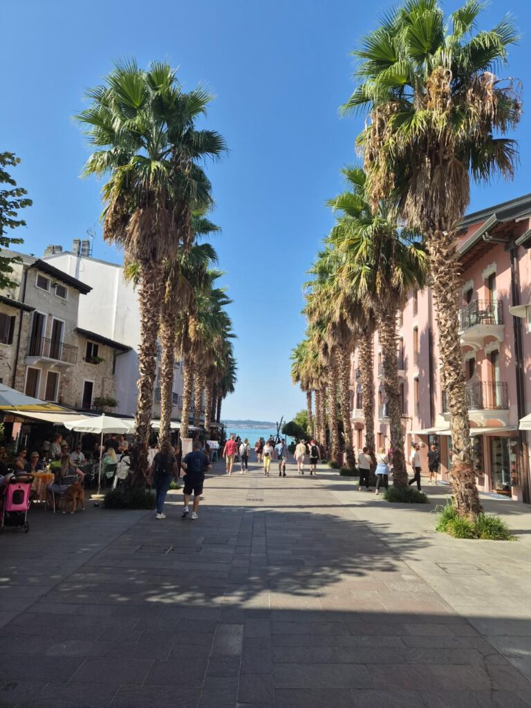 Sirmione jezero Garda, prostrana šetnica s drvoredom visokih palmi, terasama kafića i pogledom na jezero na sunčan dan.