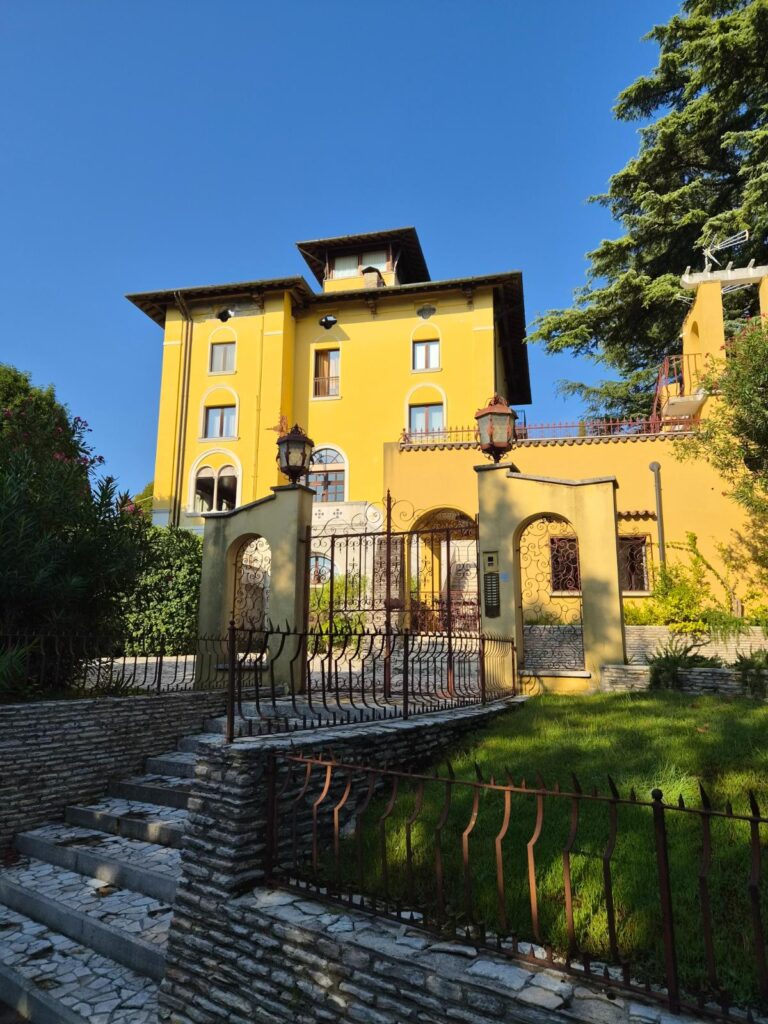 Sirmione jezero Garda, elegantna žuta vila s kovanom ogradom, kamenim stepenicama i zelenim vrtom pod vedrim plavim nebom.